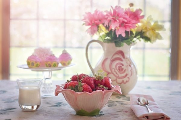 Вазочка с клубникой на столе рядом с цветочной вазой и стаканом молока